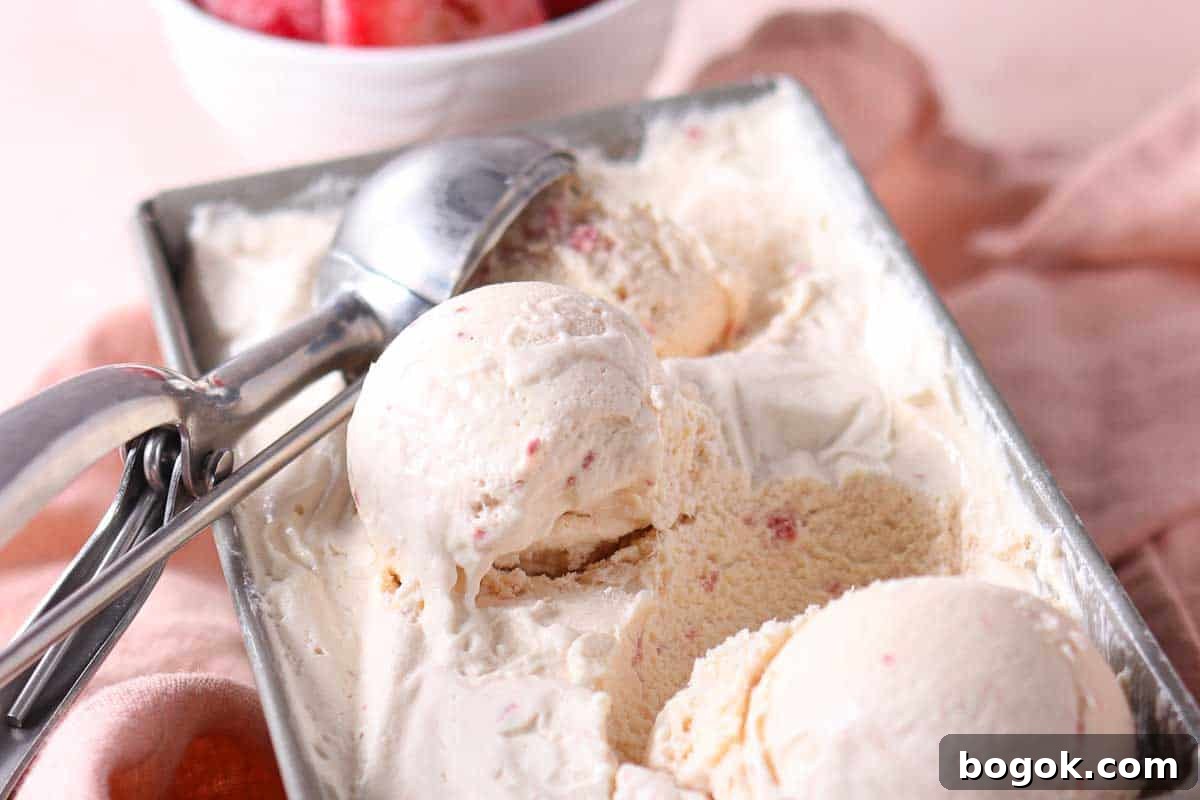 Rectangular metal container filled with scoops of creamy ice cream and a metal ice cream scoop, with a bowl of fruit in the background.