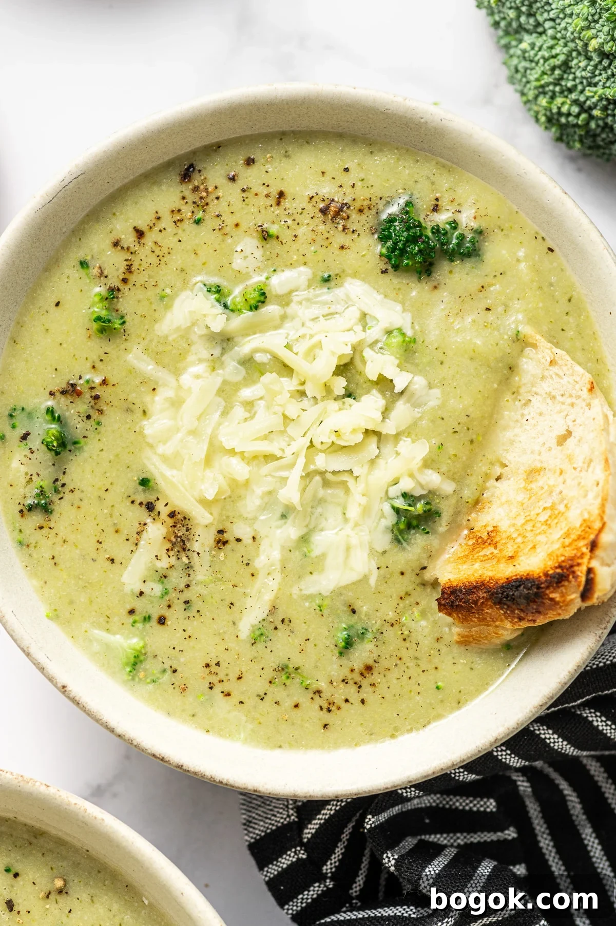 Creamy Broccoli and Cauliflower Soup Recipe for Comforting Meals 2 Overhead photo of a light beige bowl filled with creamy pale green broccoli cauliflower soup topped with shredded cheddar and cracked black pepper. A toasted bread wedge is tucked into the right side of the bowl for dipping. A second bowl is partially visible at the bottom left, with a black-and-white striped towel underneath and toast slices in the background.