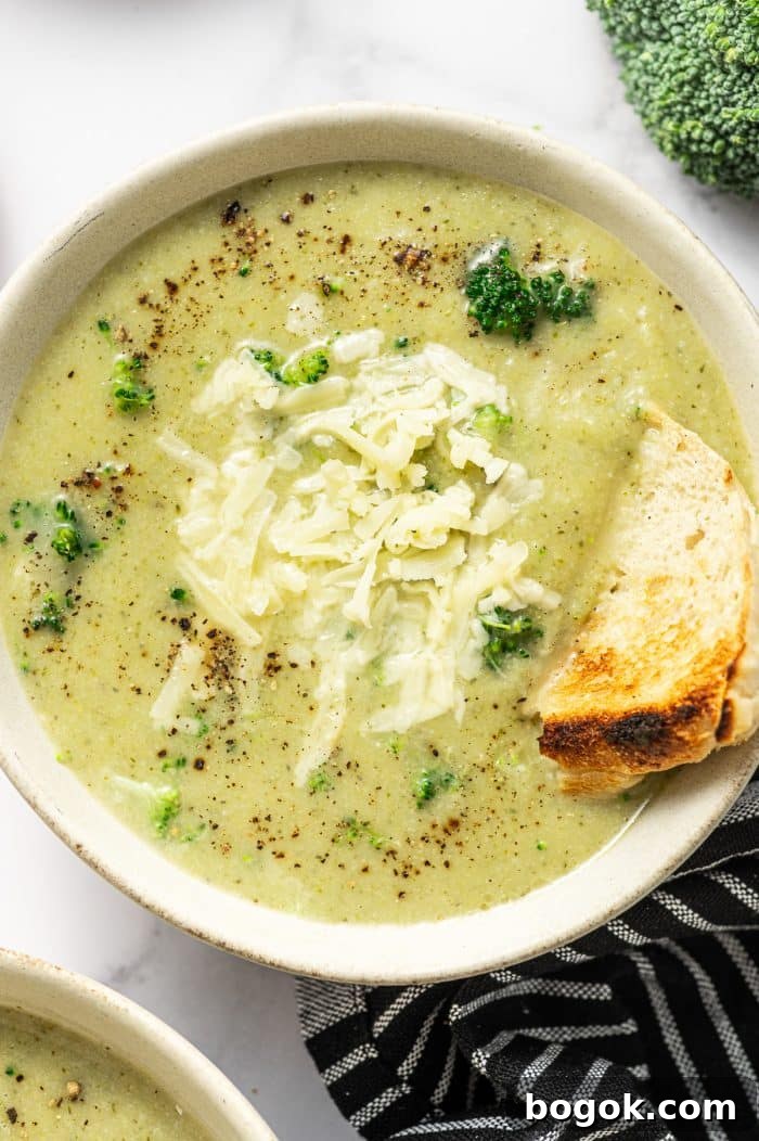 Creamy Broccoli and Cauliflower Soup Recipe for Comforting Meals 12 Overhead photo of a light beige bowl filled with creamy pale green broccoli cauliflower soup topped with shredded cheddar and cracked black pepper. A toasted bread wedge is tucked into the right side of the bowl for dipping.