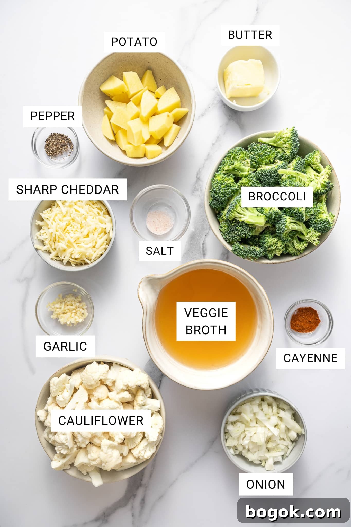Creamy Broccoli and Cauliflower Soup Recipe for Comforting Meals 3 Overhead ingredient photo on a white marble surface with text overlays labeling each item: POTATO, BUTTER, PEPPER, SHARP CHEDDAR, SALT, BROCCOLI, VEGGIE BROTH, CAYENNE, GARLIC, CAULIFLOWER, and ONION.