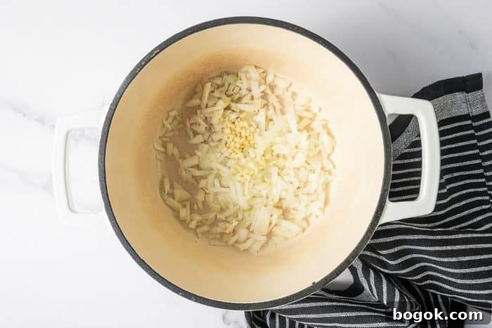 Creamy Broccoli and Cauliflower Soup Recipe for Comforting Meals 4 Overhead shot of a cream-colored enameled Dutch oven with a black rim and white handles. Chopped onion and minced garlic are sautéing in the bottom of the pot.