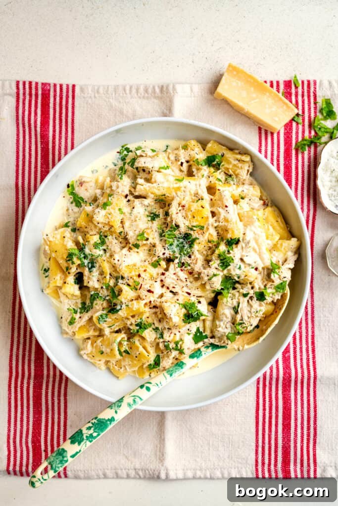 In a white bowl, Crockpot Creamy Chicken Pasta is topped with parsley and grated cheese. The bowl sits on a beige cloth with red stripes, accompanied by a cheese wedge. A floral-patterned spoon rests in the comforting ensemble.