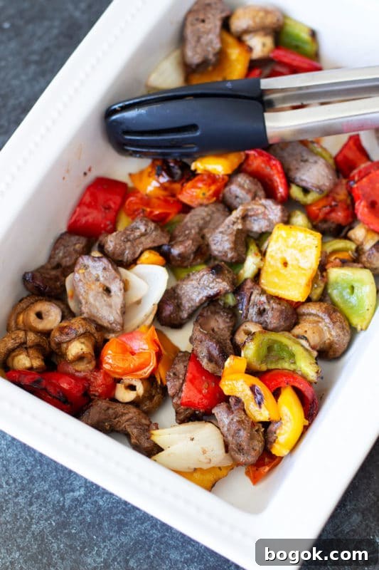 White dish containing steak and vegetable kebabs, tongs in dish.
