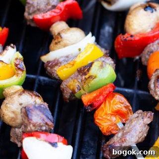 Steak and vegetables on three metal skewers on a grill.