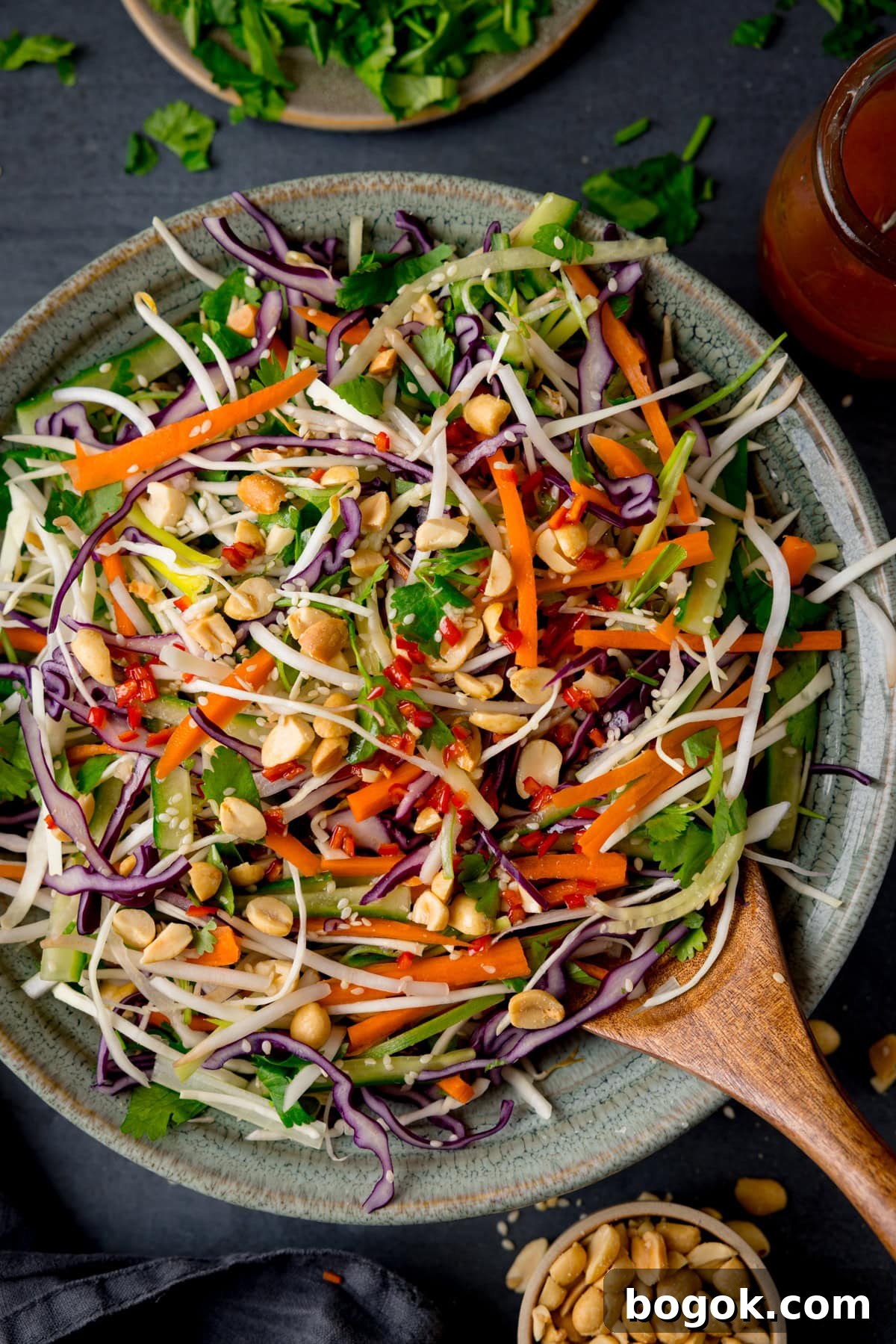 A tall shot of Asian Slaw. The Asian Slaw is in a large light grey bowl, with a wooden spoon sticking out of the dish. In the top right of the image, you can slightly see a clear glass dish with the remaining dressing in it. In the top middle of the image, you can see a grey dish with chopped coriander on it and scattered around the dish. On the bottom right of the image, you can see a light brown dish filled with peanuts and some peanuts scattered around it, there is also a slate grey napkin tucked next to it. This is all set on a dark grey background.