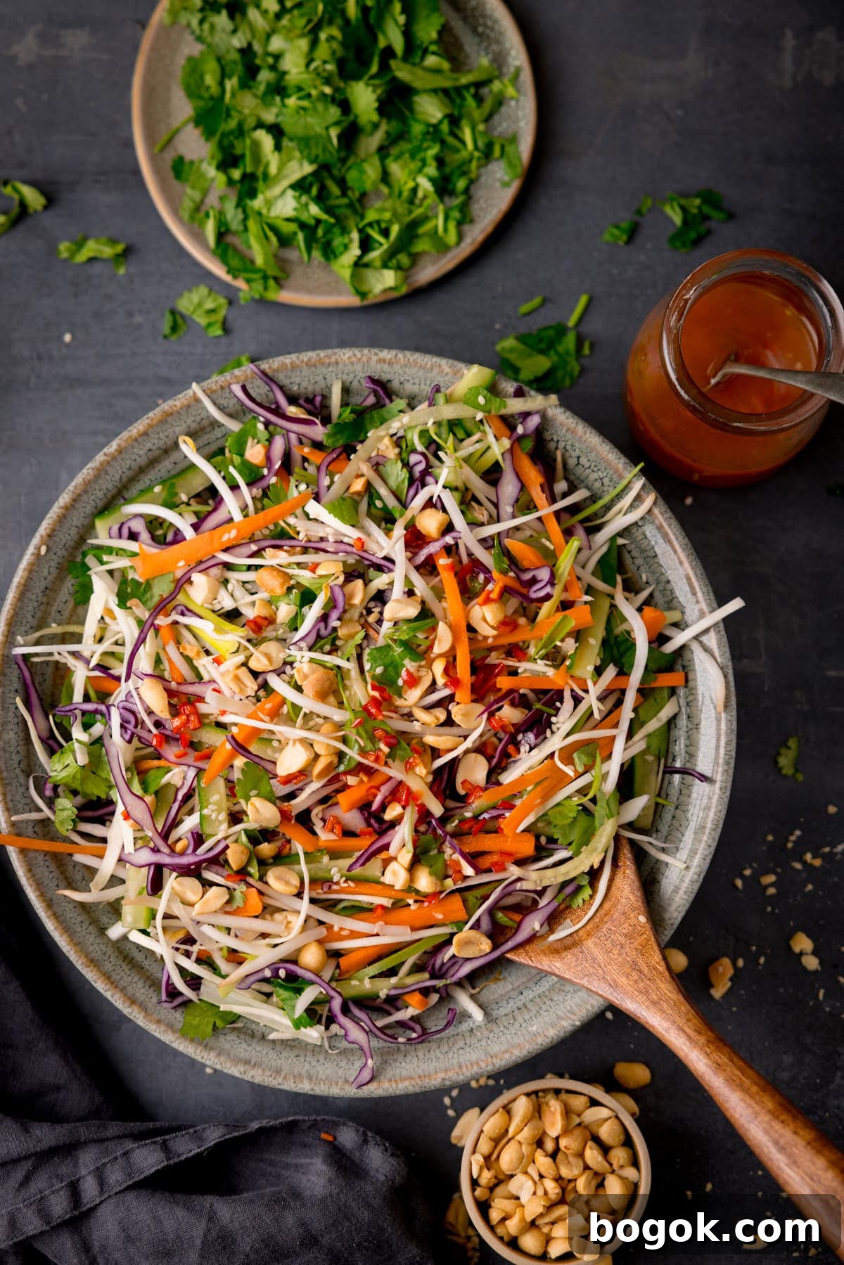 A tall, overhead shot of Asian Slaw. The Asian Slaw is in a large light grey bowl, with a wooden spoon sticking out of the dish. In the top right of the image, you can slightly see a clear glass dish with the remaining dressing in it, with a silver spoon sticking out of it. In the top middle of the image, you can see a grey dish with chopped coriander on it and scattered around the dish. On the bottom right of the image, you can see a light brown dish filled with peanuts and some peanuts scattered around it, there is also a slate grey napkin tucked next to it. This is all set on a dark grey background.
