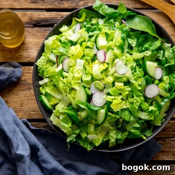Large bowl of green salad on a wooden surface. Blue napkin and jar of salad dressing is next to the bowl.