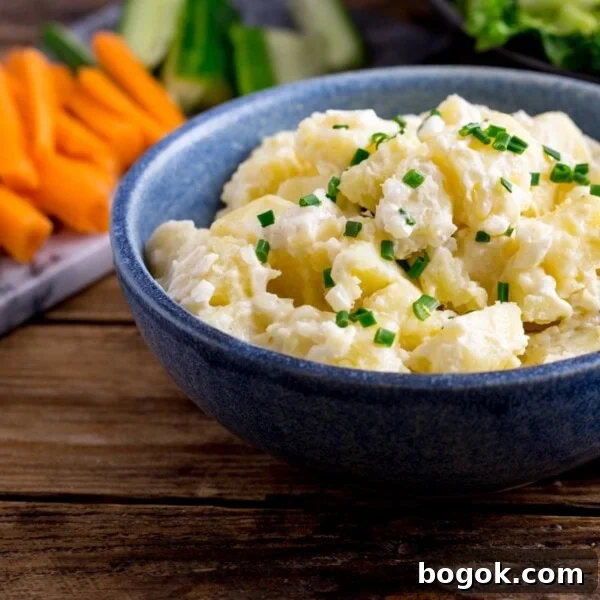 Creamy potato salad in a blue bowl on a wooden table. Chopped vegetables in the background.