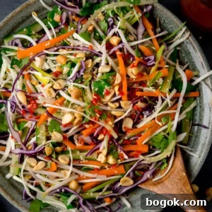 A square, closeup shot of Asian Slaw. The Asian Slaw is in a large light grey bowl, with a wooden spoon sticking out of the dish. In the top right of the image, you can slightly see a clear glass dish with the remaining dressing in it. This is all set on a dark grey background.