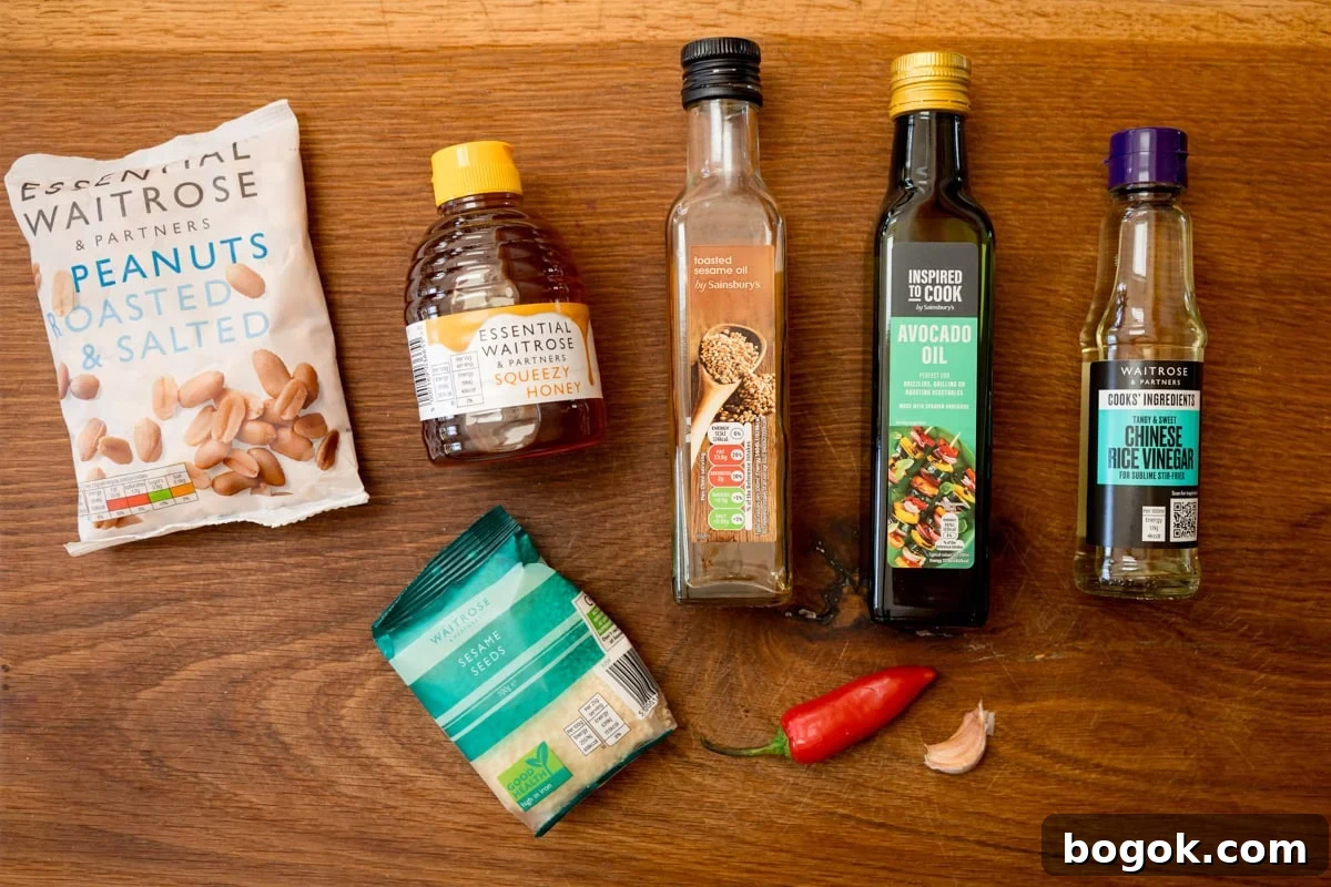 An overhead shot of the ingredients for Asian Slaw Dressing. They are laid out on a wooden cutting board. They are as follows: Roasted and Salted Peanuts, Honey, Sesame Seeds, Toasted Sesame Oil, Avocado Oil, Chinese Rice Vinegar, a red chilli, and a clove of garlic.