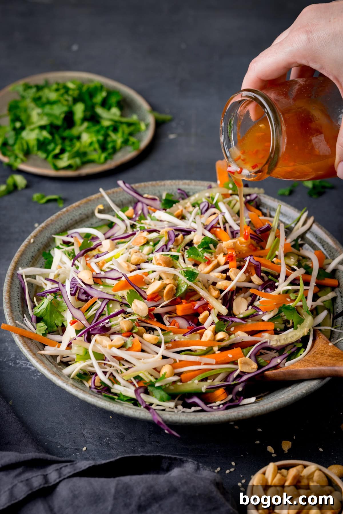 A tall shot of Asian Slaw. The Asian Slaw is in a large light grey bowl, with a wooden spoon sticking out of the dish. In the right of the image, you see a clear glass dish with the remaining dressing in it, which is being poured by a hand onto the main dish of Asian Slaw. In the top left of the image, you can see a grey dish with chopped coriander on it and scattered around the dish. On the bottom right of the image, you can see a light brown dish filled with peanuts and some peanuts scattered around it, there is also a slate grey napkin tucked next to it. This is all set on a dark grey background.