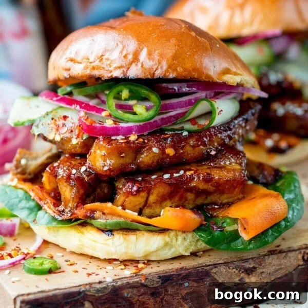 Square side-on image of toasted brioche roll filled with sticky pork belly strips and pickled carrot, red onion and cucumber strips. Topped with jalapeno slices. Served on a wooden board.