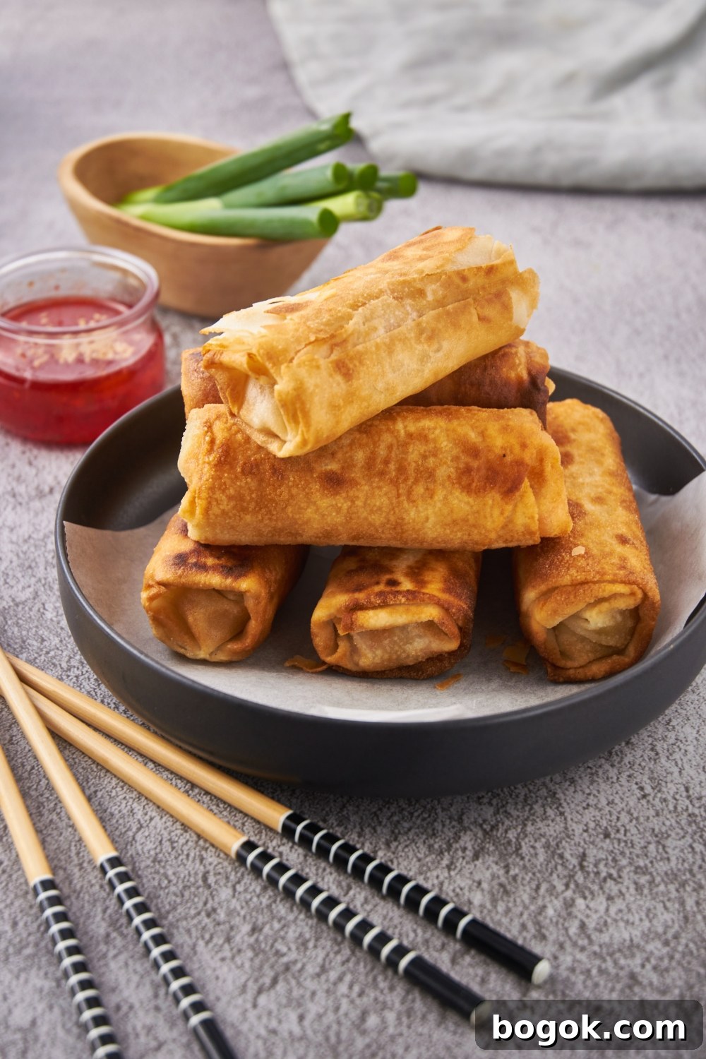 A pile of fried egg rolls stacked on top of each other on a black plate with a layer of parchment paper on the bottom.
