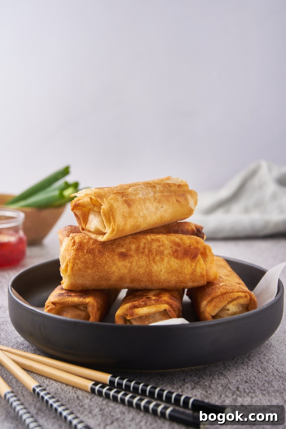 A pile of fried egg rolls stacked on top of each other on a black plate with a layer of parchment paper on the bottom.