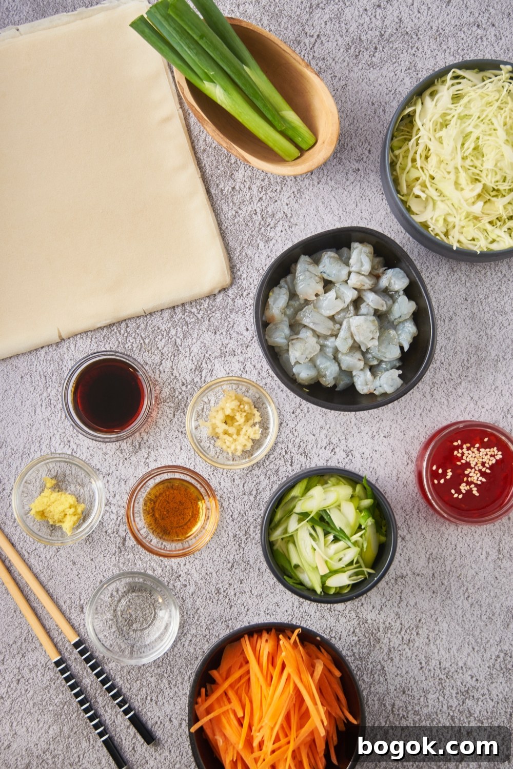 Overhead view of an assortment of ingredients including bowls of raw shrimp, shredded carrots, shredded cabbage, minced garlic, minced ginger, sweet and sour sauce, and soy sauce.