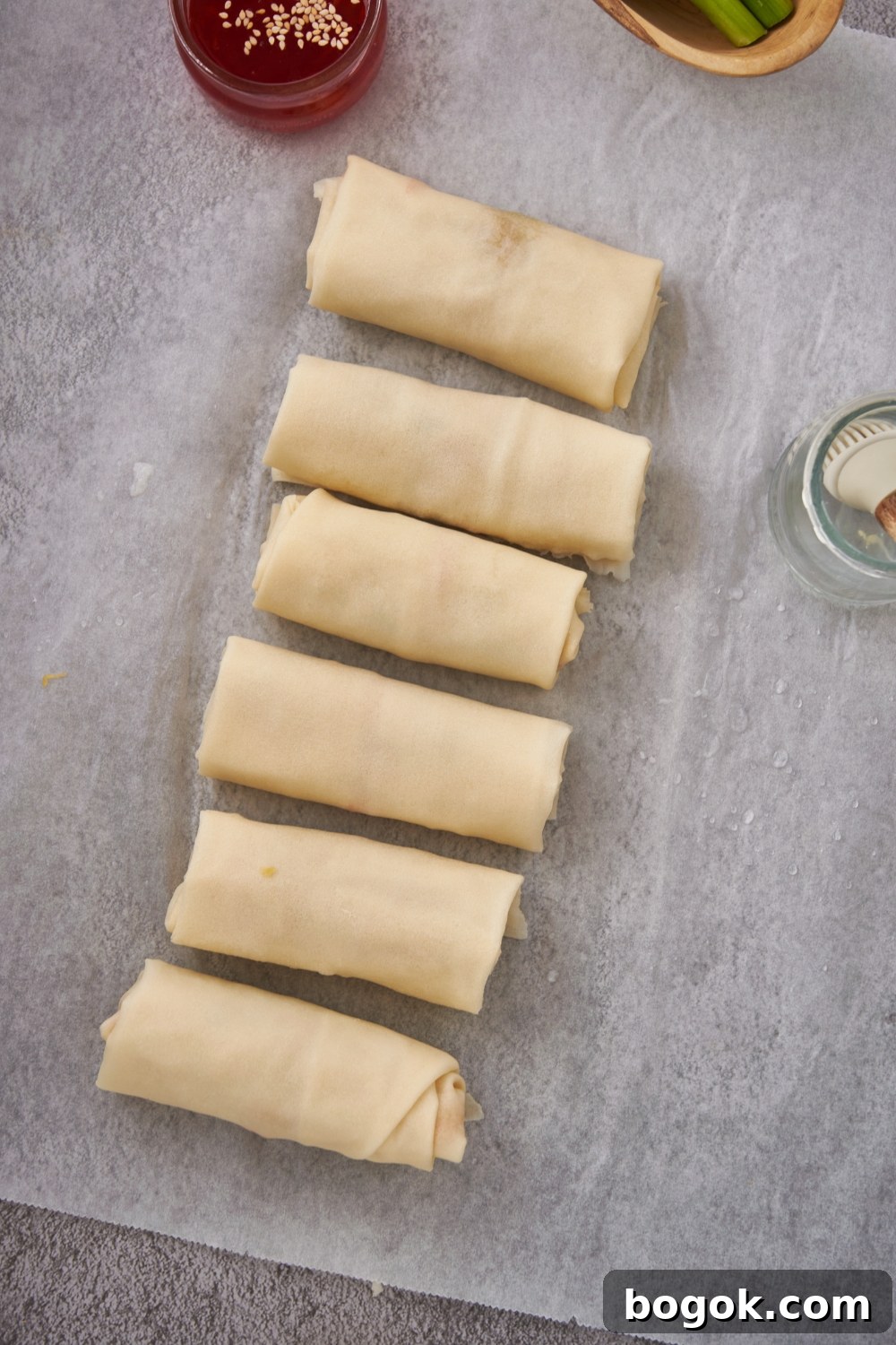 Six raw egg rolls lined up next to each other on a piece of parchment paper.