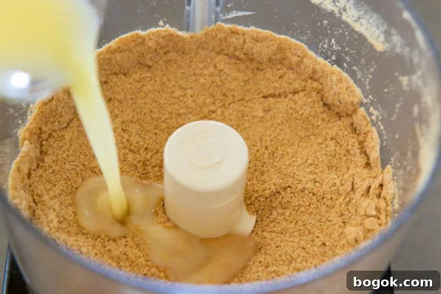 Graham Cracker Crumbs in Food Processor Bowl with Melted Butter Pouring In