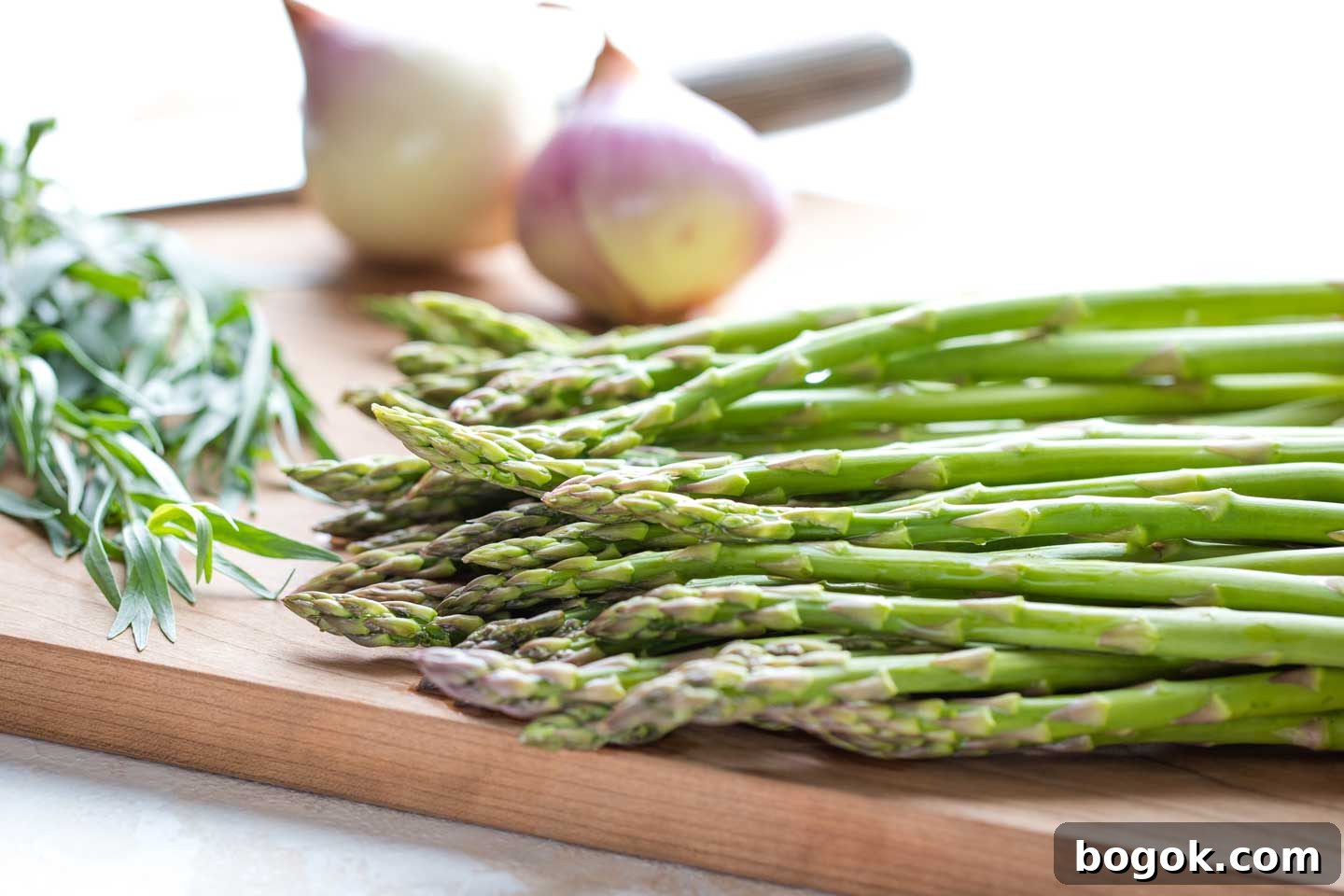 Closeup side view of fresh asparagus spears with herbs, a knife and other vegetables in the background.