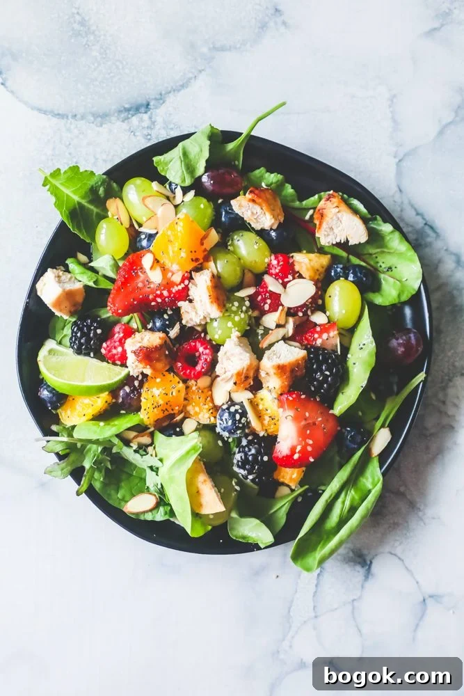 Picture of spinach salad with grapes, strawberries, blackberries, blueberries, raspberries, chicken, poppy seeds, hemp hearts, and a citrus poppyseed dressing on a black plate. 