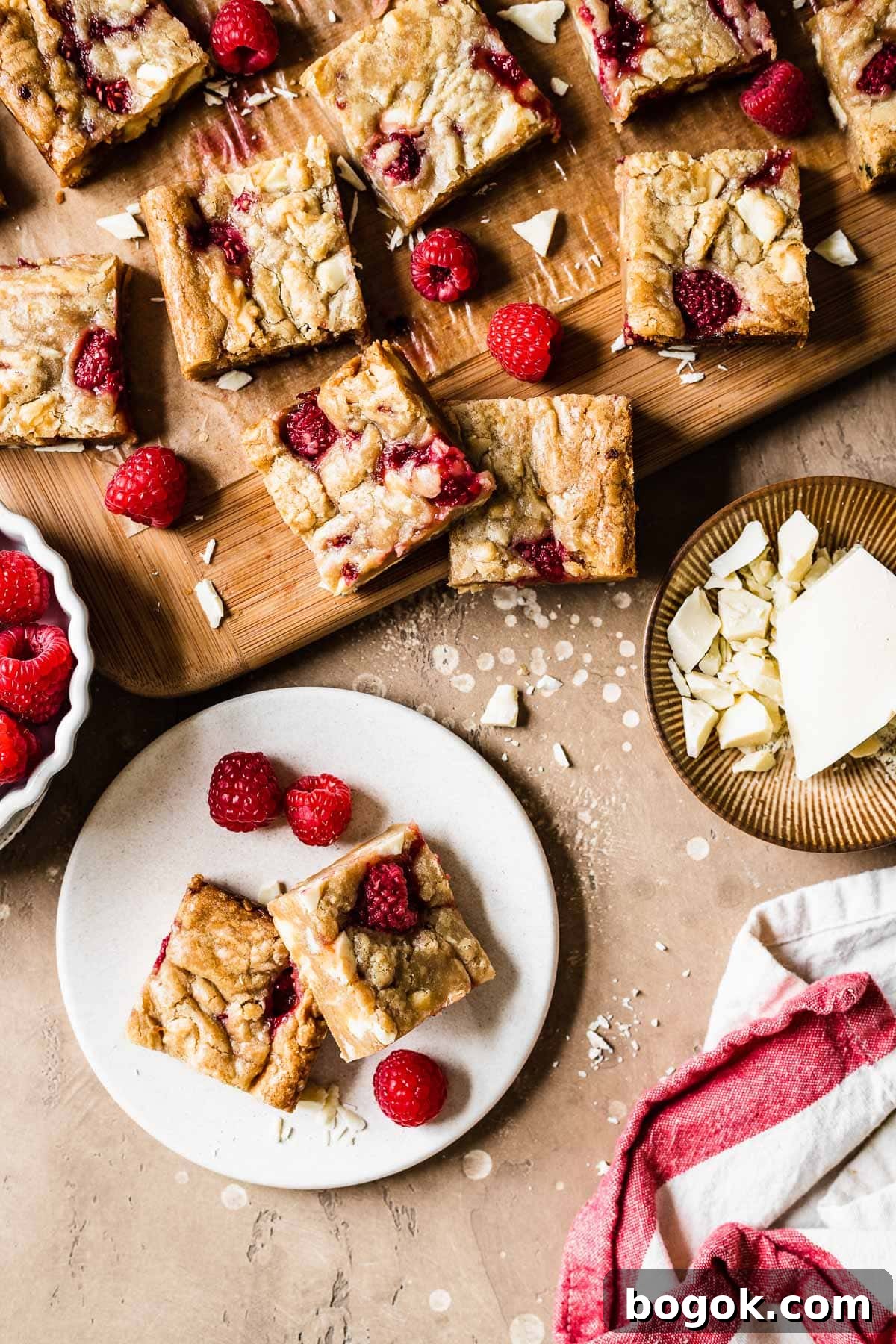 Raspberry White Chocolate Blondies Recipe with Crispy Edges 11 Two blondie squares on a small plate with raspberries and more blondie squares nearby.
