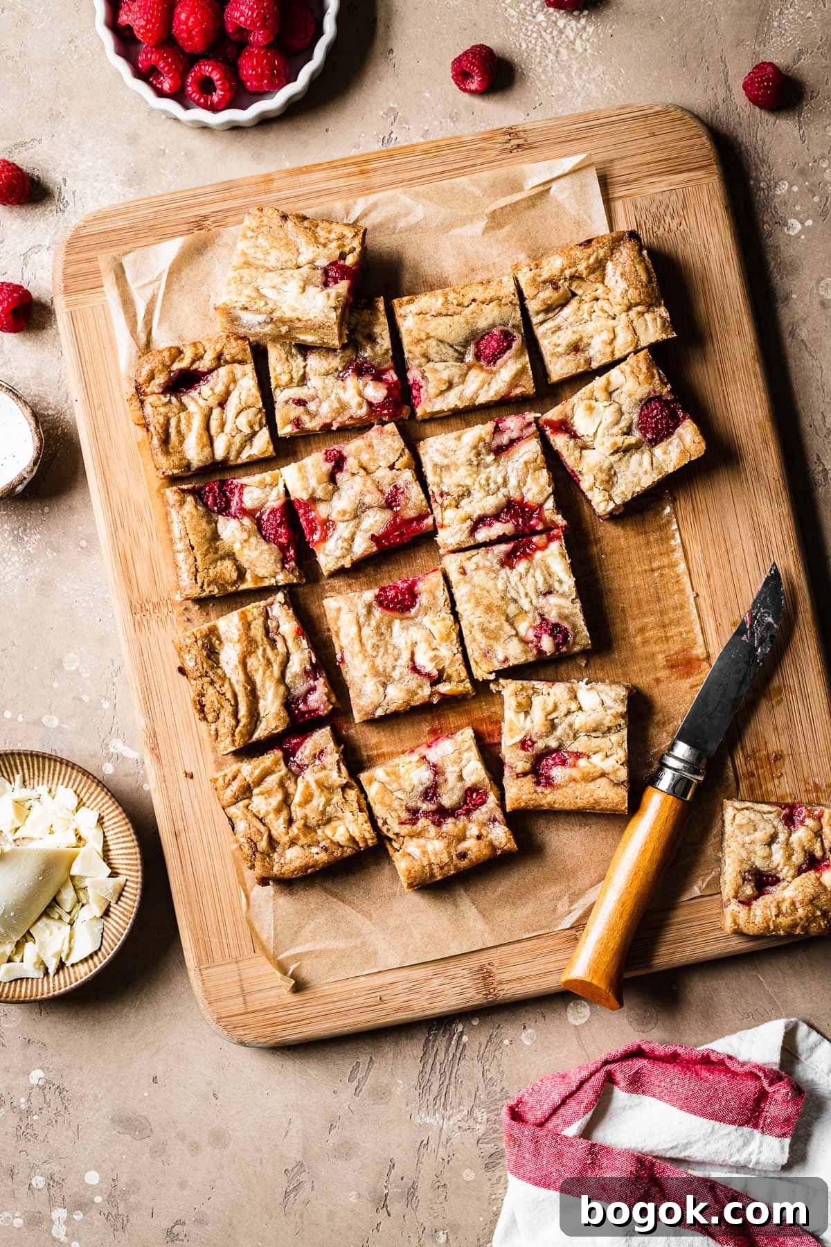 Raspberry White Chocolate Blondies Recipe with Crispy Edges 10 Raspberry and white chocolate blondies cut into squares on a wooden cutting board.