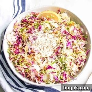 Large white bowl with brussels sprouts slaw: colorful red cabbage, shredded brussels sprouts, various nuts and seeds, lemon wedge and a creamy vegan dressing.