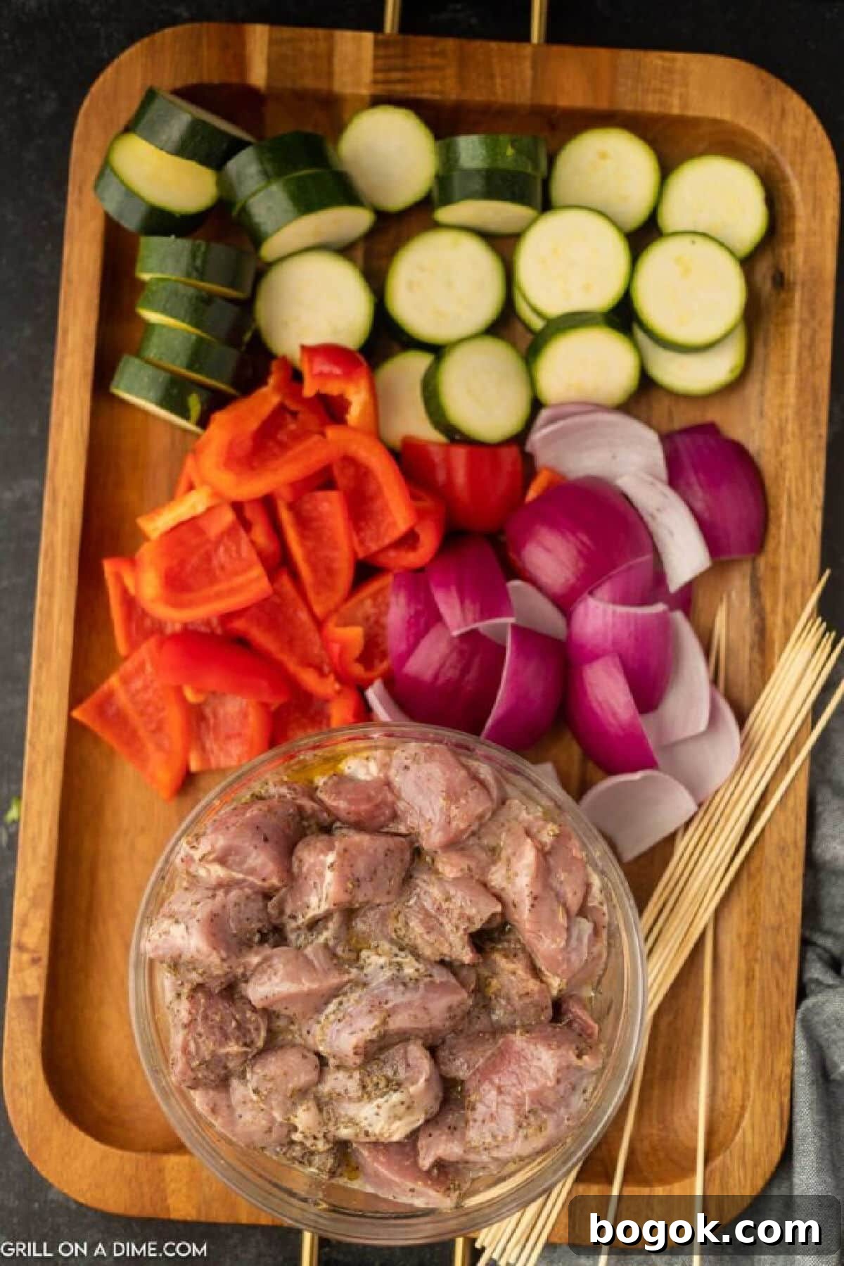 Sliced zucchini, bell peppers, and red onions on a cutting board with a bowl of marinated pork pieces with wooden skewers on the side