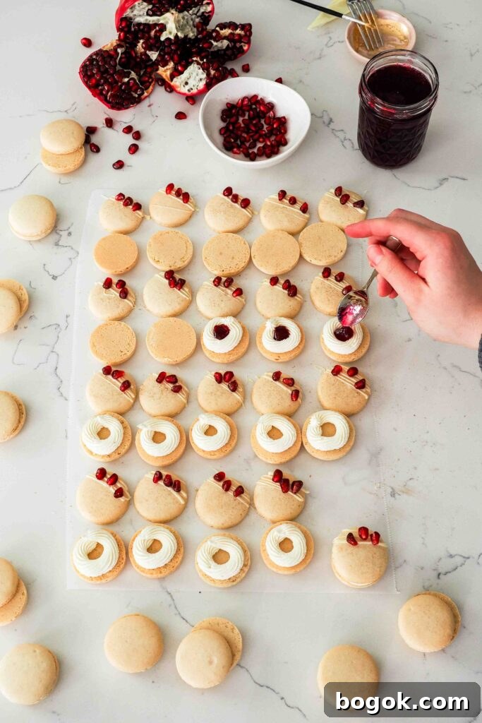 White Chocolate and Pomegranate Macarons — Recipe & Tips 19 A hand spoons a teaspoon of pomegranate jelly into the center of a white chocolate buttercream ring on a macaron.