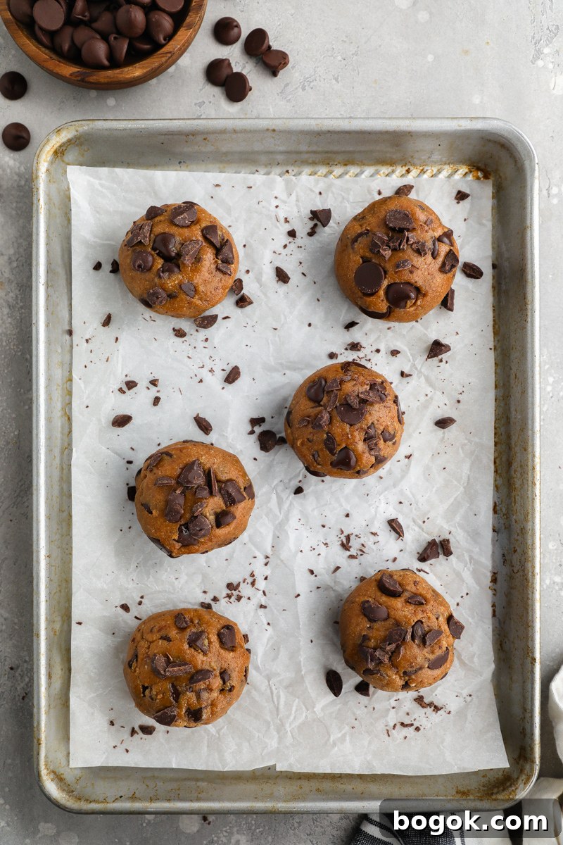 Raw chocolate chip cookie dough balls stuffed with sweetened cream cheese sitting on a baking pan next to a bowl of dark chocolate chips
