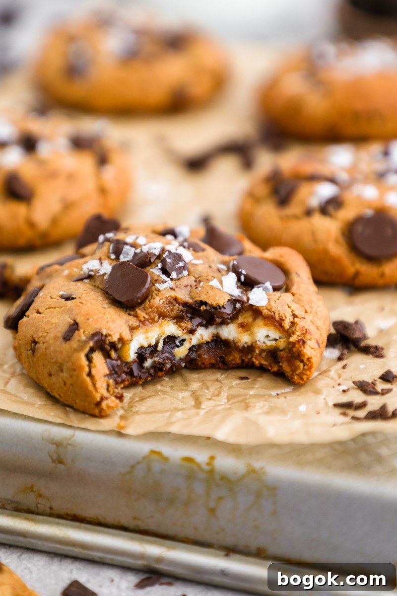 A stuffed chocolate chip cookie sitting on a baking pan with a bite taken out of it exposing it's cheesecake filling