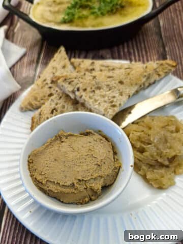 A small white dish of duck liver pate on a white plate with a spoonful of onion marmalade on the side and a few pieces of crusty brown toast.