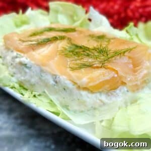 Close up of a salmon and dill terrine showing the creamy salmon layer topped with smoked salmon and dill. The terrine is arranged on lettuce leaves on a white platter.