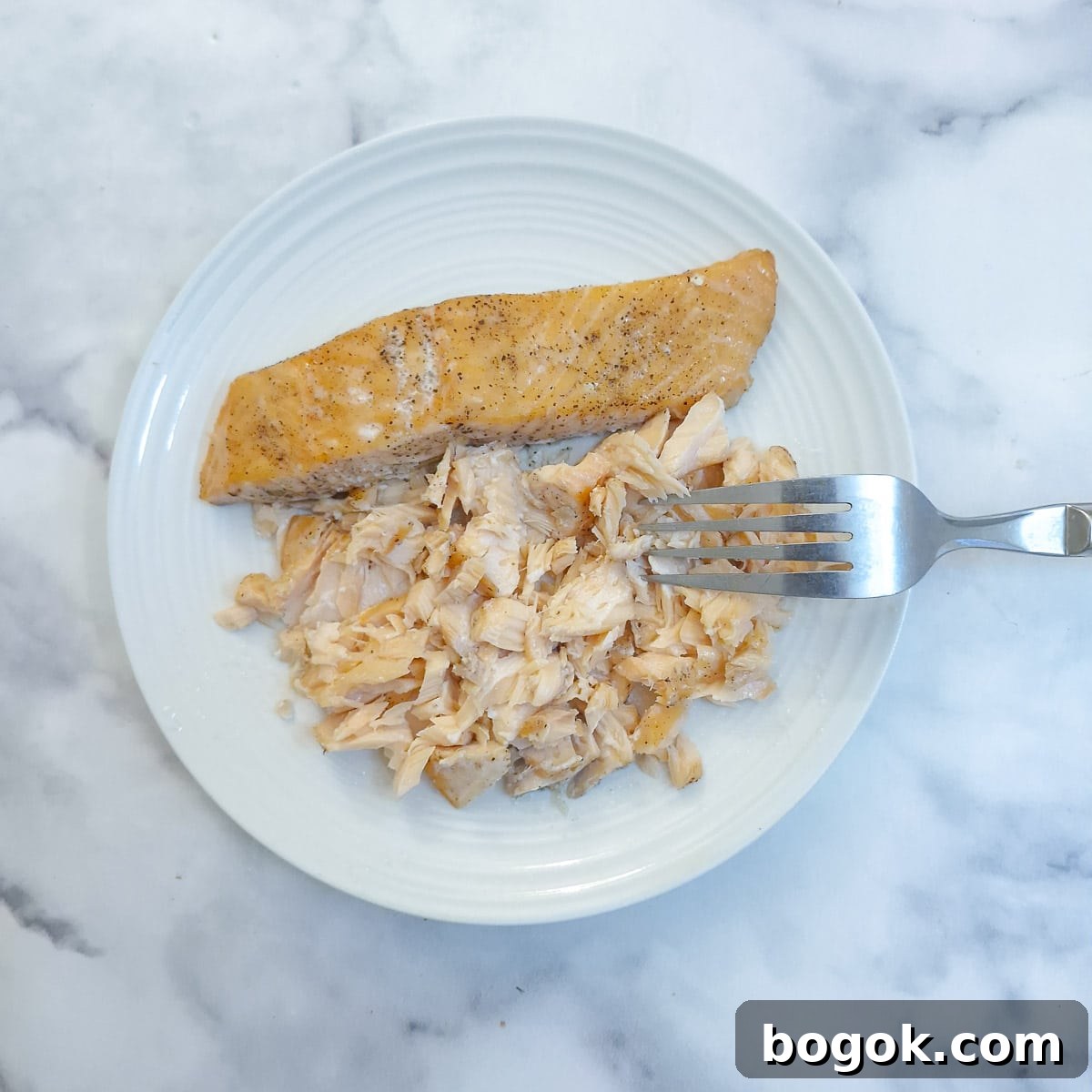 Cooked salmon being flaked with a fork on a white plate.