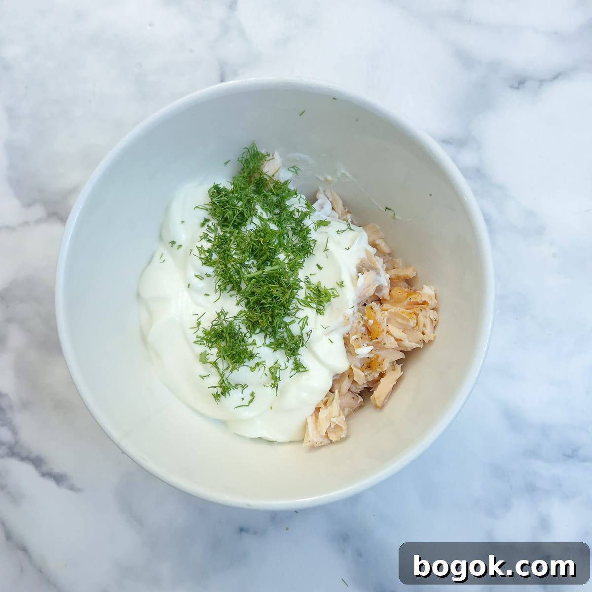 Flaked salmon in a white mixing bowl with mayonnaise, creme fraiche and chopped dill.