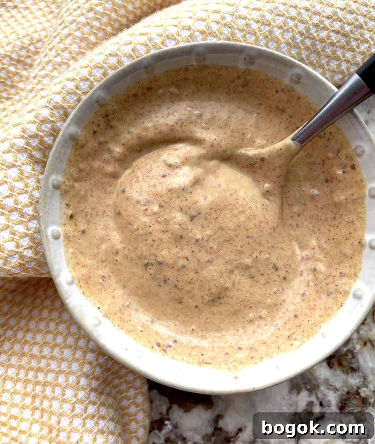 A small bowl of Creole remoulade sauce with a spoon on a marble countertop with a yellow towel.