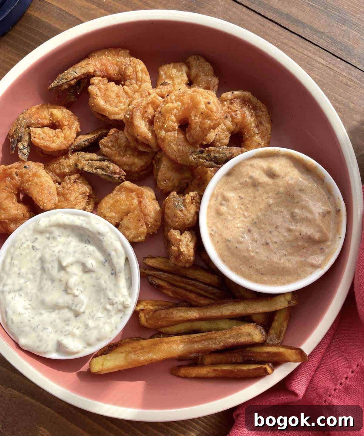 A pink plate with fried shrimp, tartar sauce, remoulade sauce, and french fries on it.