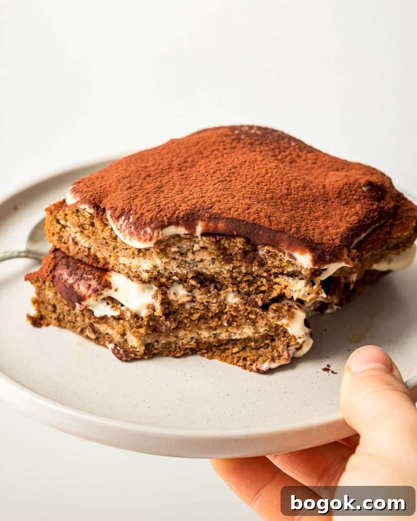 A hand holding a grey plate with a single serve tiramisu with a bite missing.