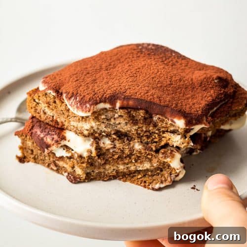 A hand holding a grey plate with a single serve tiramisu with a bite missing.