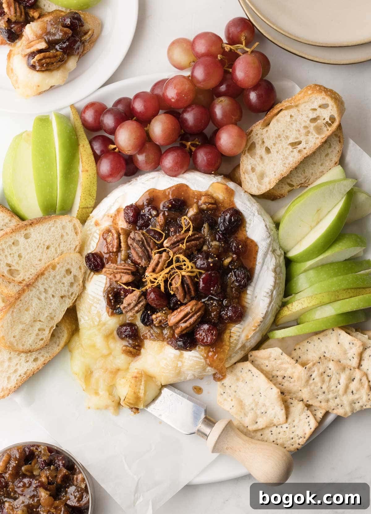 A platter of bake brie cheese with fig jam, cranberries, pecans, and bread with apple slices.