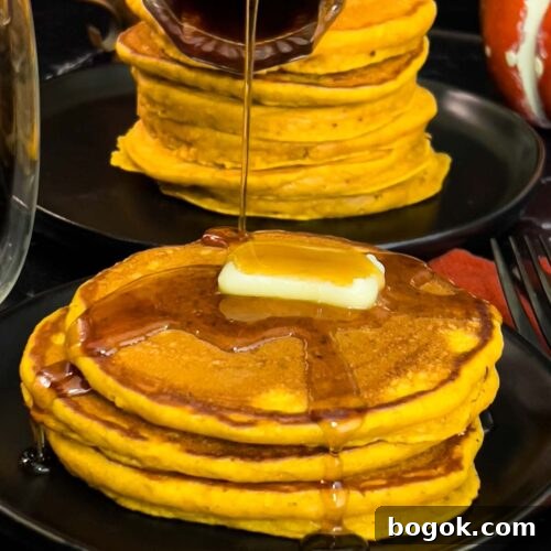 Stack of fluffy pumpkin pancakes with butter and maple syrup, golden edges on a dark plate.
