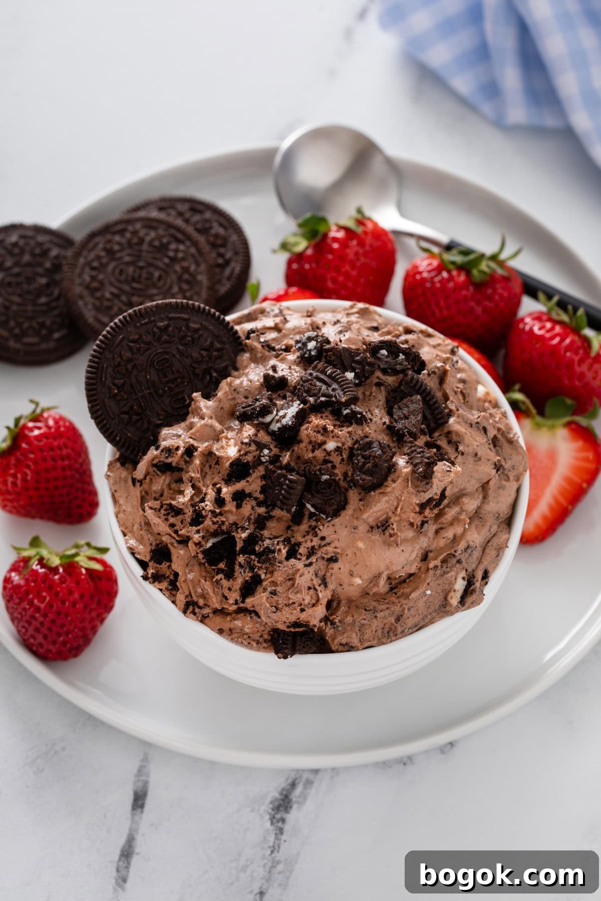 Small white bowl filled with oreo fluff set on a white plate with oreo cookies and fresh strawberries.