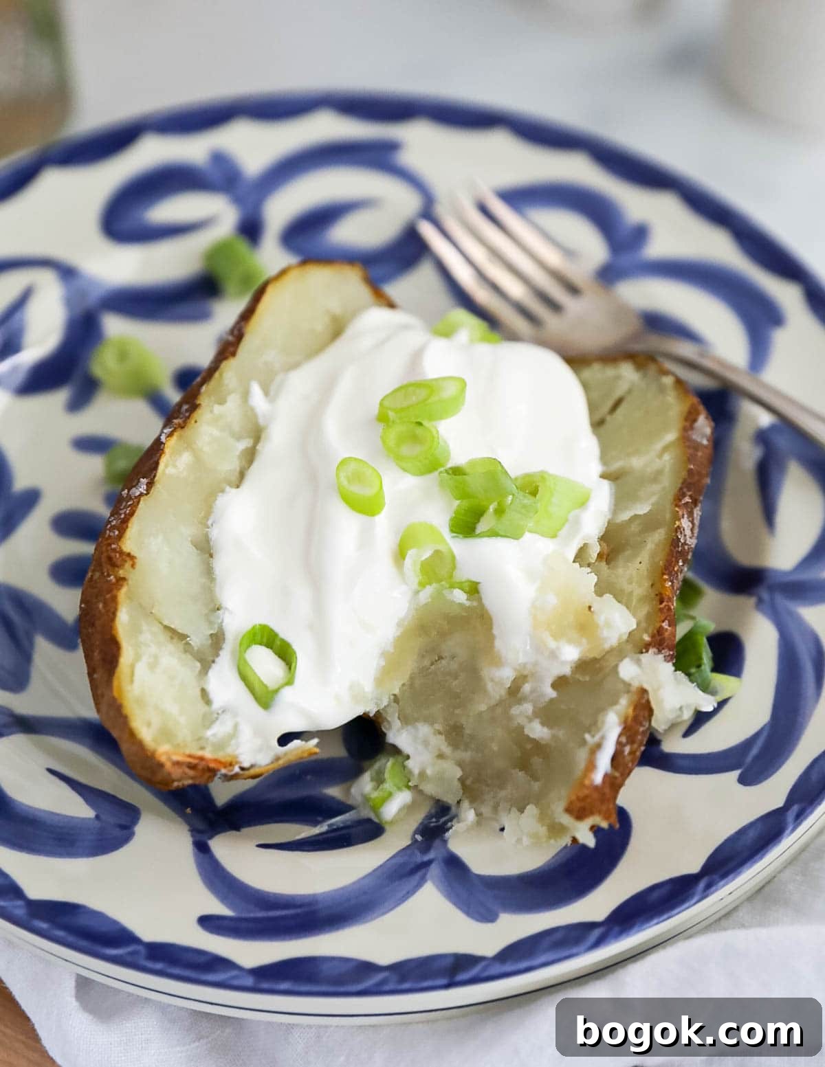 A baked potato on a blue and white plate. The potato is sliced in half, there is a dollop of sour cream and sliced scallions inside the potato, and a fork next to the potato.