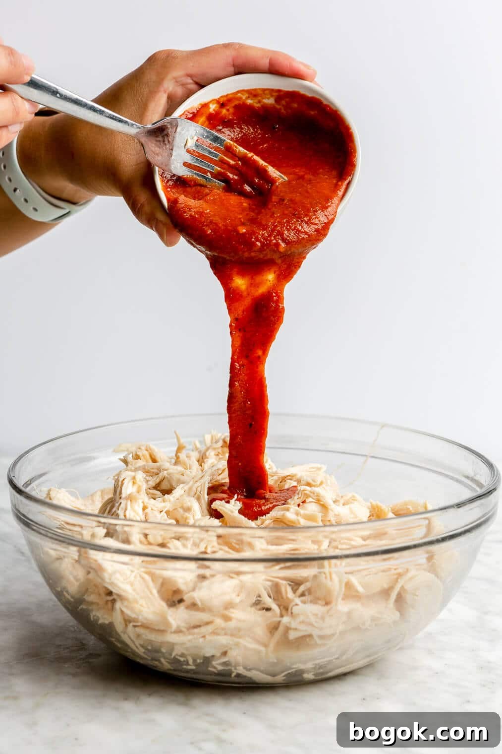 BBQ Chicken and Sweet Potato Casserole Recipe 6 A person pouring BBQ sauce over a glass bowl of shredded chicken.