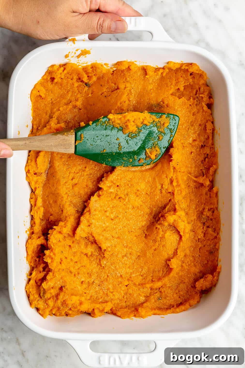 BBQ Chicken and Sweet Potato Casserole Recipe 7 A person using a rubber spatula to spread mashed sweet potato in a white casserole dish.