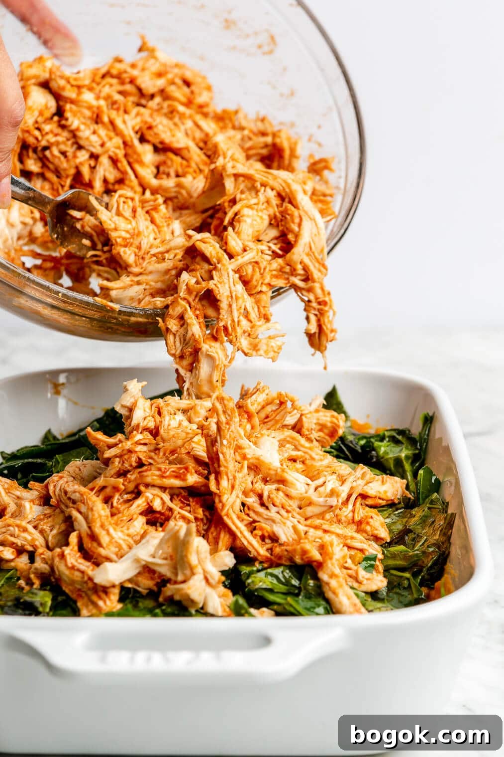 BBQ Chicken and Sweet Potato Casserole Recipe 9 A person spooning shredded BBQ chicken over sweet potato and kale in a casserole dish.
