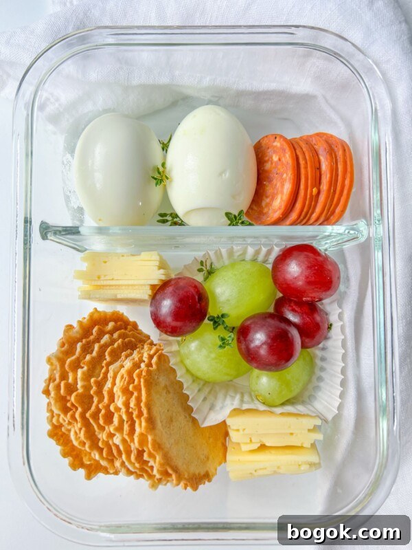 A glass container holds an Adult Lunchable with hard-boiled eggs, sliced pepperoni, cheese slices, crackers, and red and green grapes, all separated into neat sections on a white background.