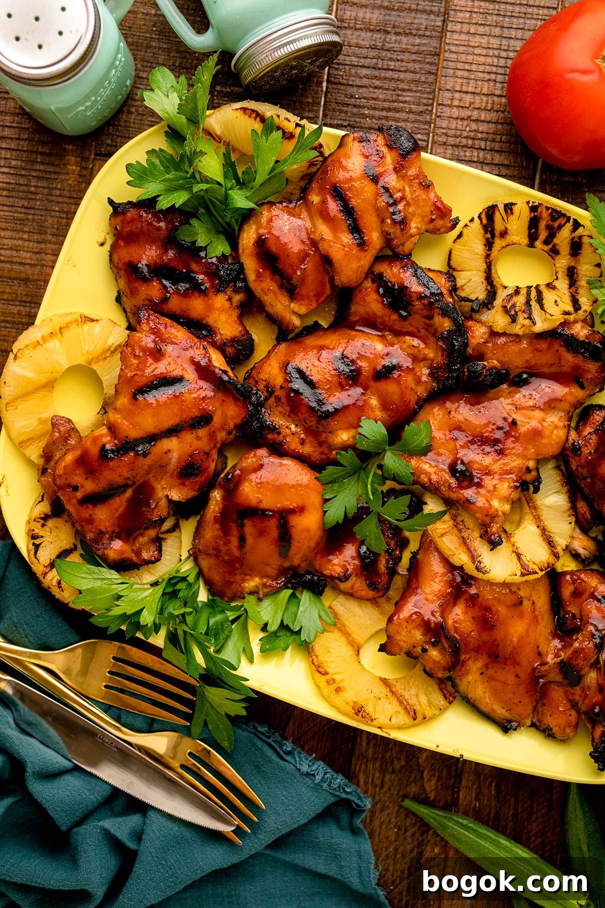Hawaiian-Style BBQ Chicken with Pineapple Glaze 11 Grilled Hawaiian BBQ chicken thighs with perfect grill marks rest on a yellow platter, surrounded by juicy grilled pineapple slices and garnished with fresh parsley. Forks and a teal napkin complete the inviting setup.