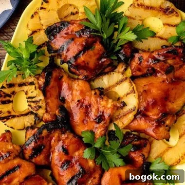 Hawaiian-Style BBQ Chicken with Pineapple Glaze 12 Grilled Hawaiian BBQ chicken thighs and pineapple rings, garnished with parsley, rest on a yellow platter, surrounded by fresh tomato, lettuce, and pineapple.