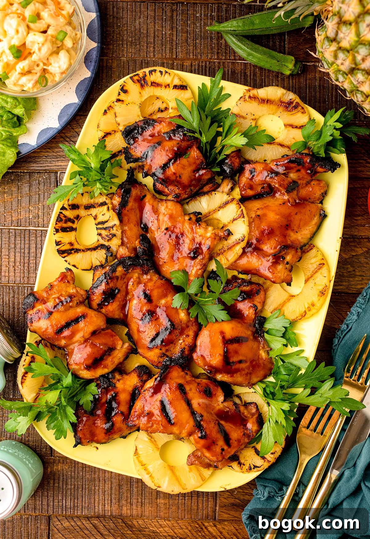 Hawaiian-Style BBQ Chicken with Pineapple Glaze 3 Grilled Hawaiian BBQ chicken thighs with perfect grill marks are served on a plate, garnished with grilled pineapple slices and fresh parsley, surrounded by cutlery and a green napkin.