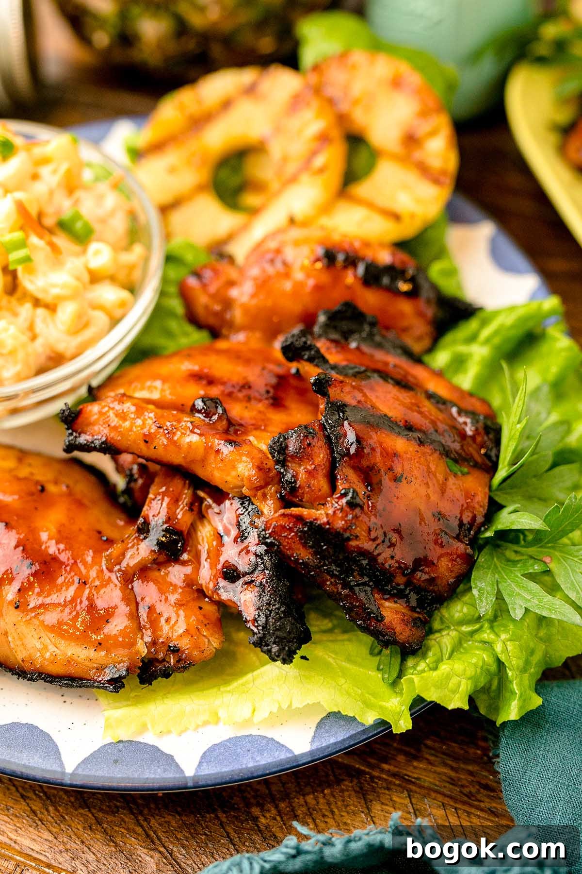 Hawaiian-Style BBQ Chicken with Pineapple Glaze 6 Grilled Hawaiian BBQ chicken thighs with a charred glaze are served on crisp lettuce alongside grilled pineapple slices and a side of creamy pasta salad, all arranged beautifully on a plate.